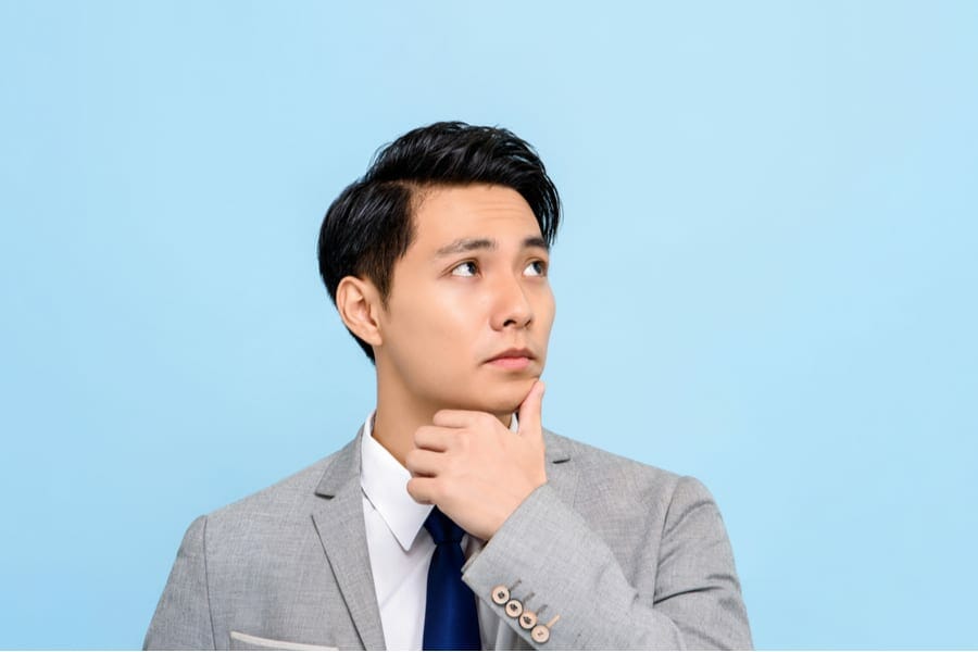 Panoramic portrait of serious young Asian businessman thinking with hand on chin isolated blue background with copy space