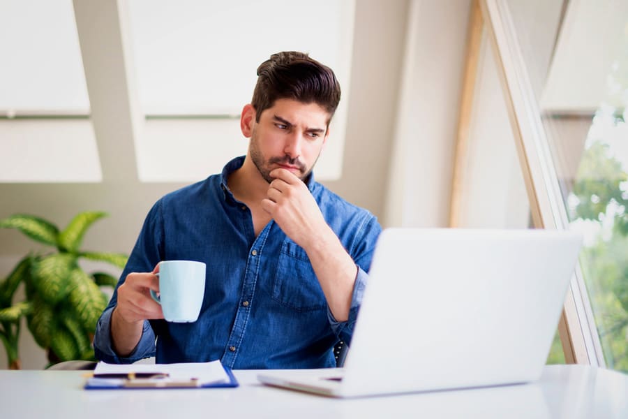Businessman looking thoughtfully while drinking tea and working on laptop at home. Home office.