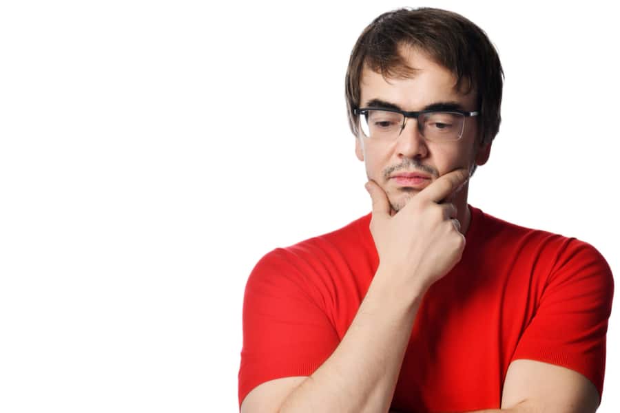 Portrait of pensive unshaved adult man in glasses and red t-shirt touching chin and thinking over white background wit free copy space. Expressing emotions and moods concept
