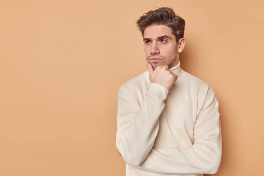 Indoor shot of handsome man holds chin looks away pensively concentrated away considers future plans dressed in casual turtleneck isolated over beige background with copy space for your text