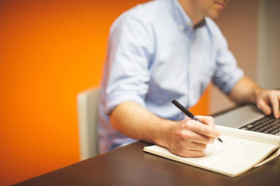 man writing notes while using laptop