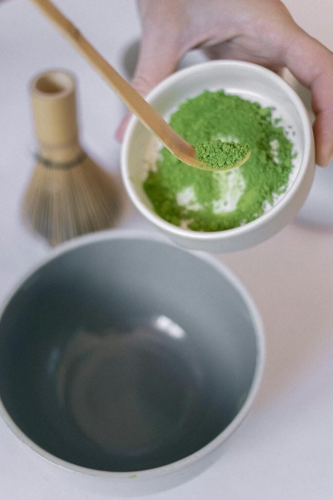 A hand scoops kratom powder into a bowl next to a bamboo whisk