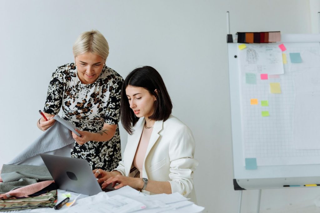 Two women work together at a desk covered with fabric samples and notes, strategizing sustainable growth for fashion brands
