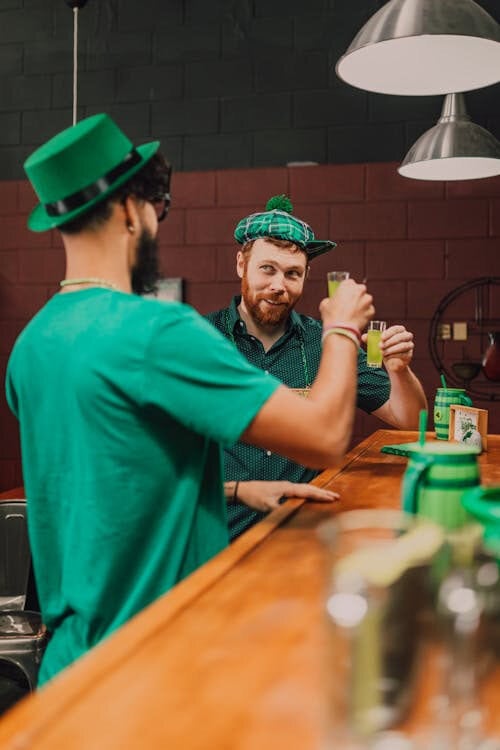 Two men in green festive outfits clink glasses at a bar, celebrating St. Patrick’s Day with trusted quality drinks